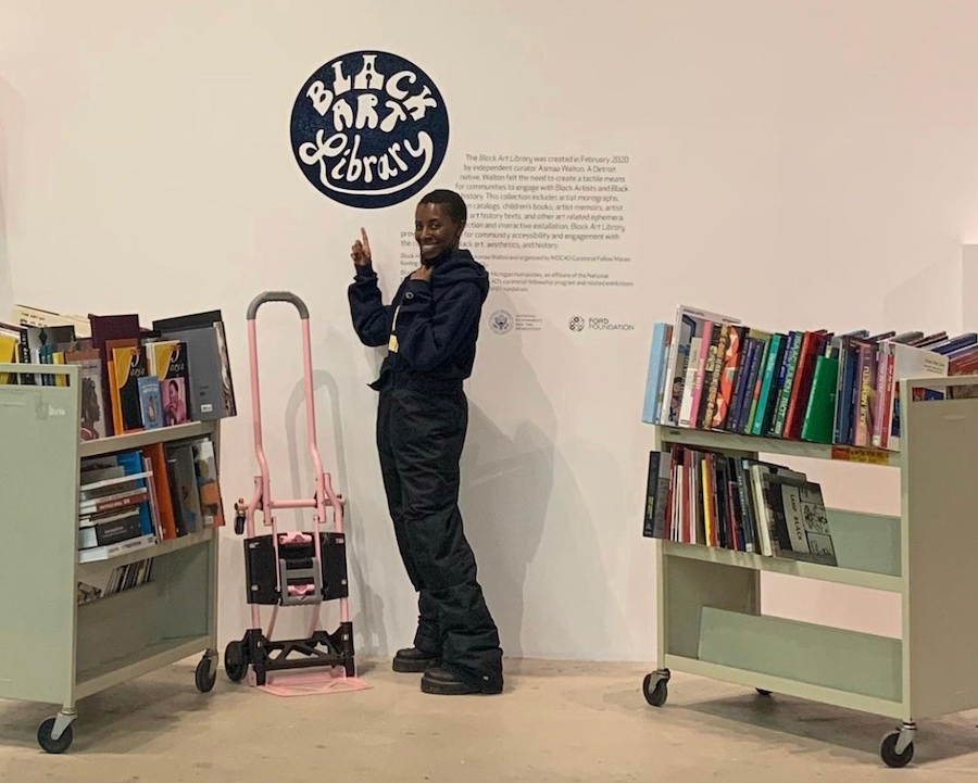 Black woman stands between two green book carts with a pink hand truck in front of wall text reading "Black Art Library" and describing the exhibition